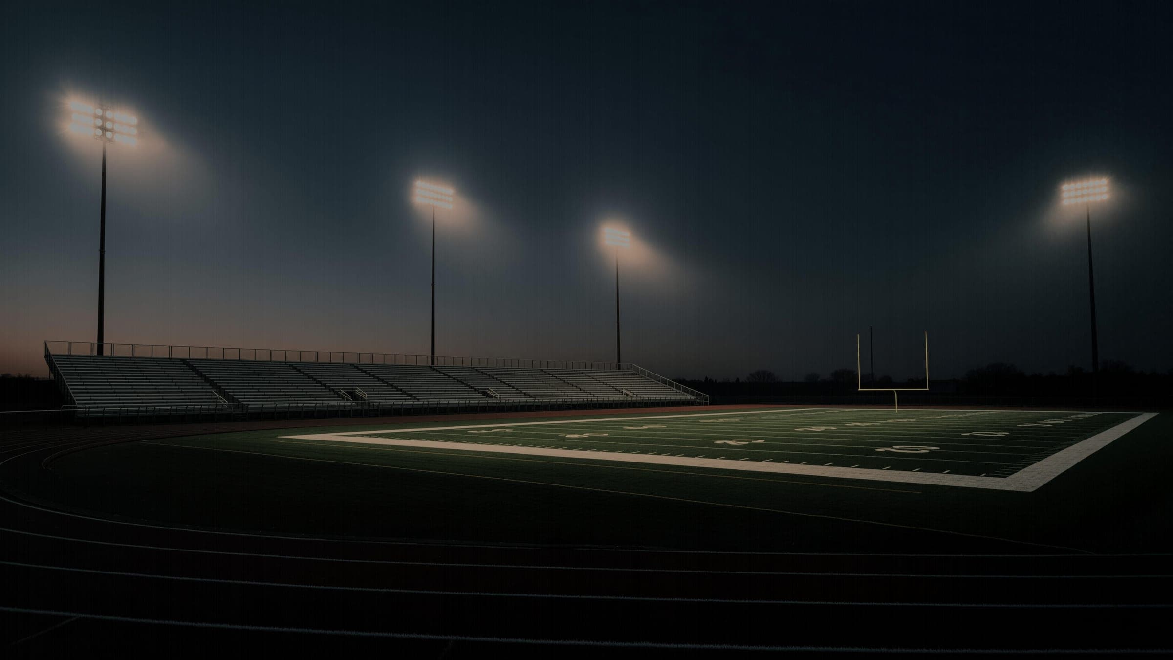 An empty high school football stadium at dusk, floodlight towers burning through haze over an unlit field and empty bleachers.