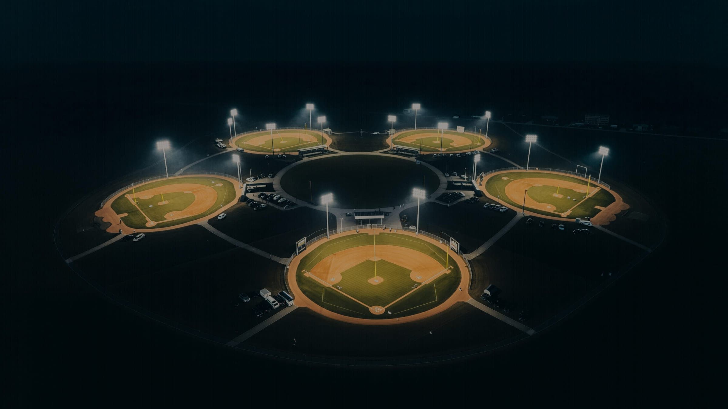An aerial view at night of a tournament complex, six lit baseball diamonds arranged in a cloverleaf against surrounding darkness.